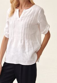 Donna che indossa una blusa in lino bianca, con vestibilità ampia, pieghe verticali e scollatura a V, abbinata a pantaloni neri, con una mano in tasca.