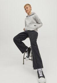 Poiss, kes kannab heledat halli kapuutsiga pusa, musti laiade säärtega teksapükse ja musti kõrge säärega tossusid, istub turutoolil ühevärvilise tausta ees.