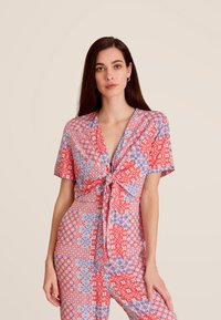 Combinaison à motifs rouge et bleu avec des manches courtes et un détail noué à l'avant. Tissu léger arborant des motifs géométriques.