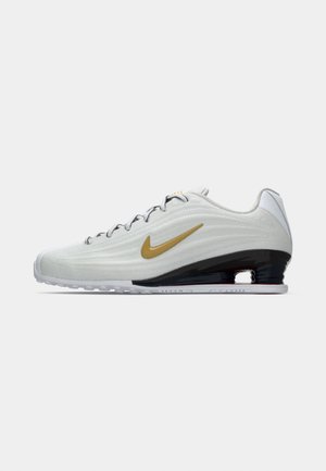 Zapatilla Nike de perfil bajo en color blanco con el logo de Swoosh dorado, suela intermedia negra con amortiguación de aire y cordones grises, mostrada en vista lateral.