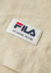 La pièce en tissu beige présente le logo « FILA » en rouge et bleu marine, avec des détails numériques en dessous. Les bords sont cousus et la texture est lisse.
