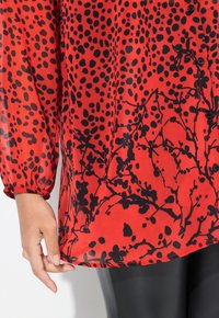 Blusa roja ligera con lunares negros y patrones florales, con un cierre frontal de botones y mangas elásticas. Textura de tela suave.