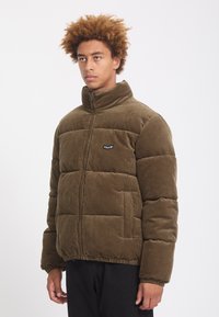 Jeune homme aux cheveux bouclés portant une doudoune en velours côtelé marron avec un col montant et un pantalon noir sur fond blanc.