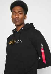 Haut à capuche noir en mélange de coton, avec une poche avant, une capuche avec cordon de serrage et un logo doré. Poche latérale avec zip et détail de tag rouge.