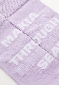 Purple socks featuring white block text stating "MAKIA COMPANY THROUGH THE ROUGH SEAS." Soft knit material with a textured finish.