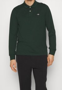 Long-sleeve green polo shirt made of cotton, featuring a collar, two-button placket, and a small embroidered logo on the chest.