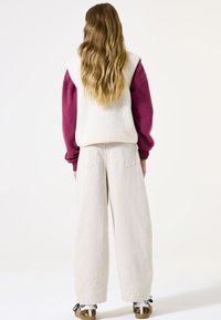 Fleece-Weste in Weiß über einem burgunderfarbenen Sweatshirt, kombiniert mit lockeren beigen Hosen. Auffällige Details sind die strukturierte Oberfläche und der weit geschnittene Sitz.