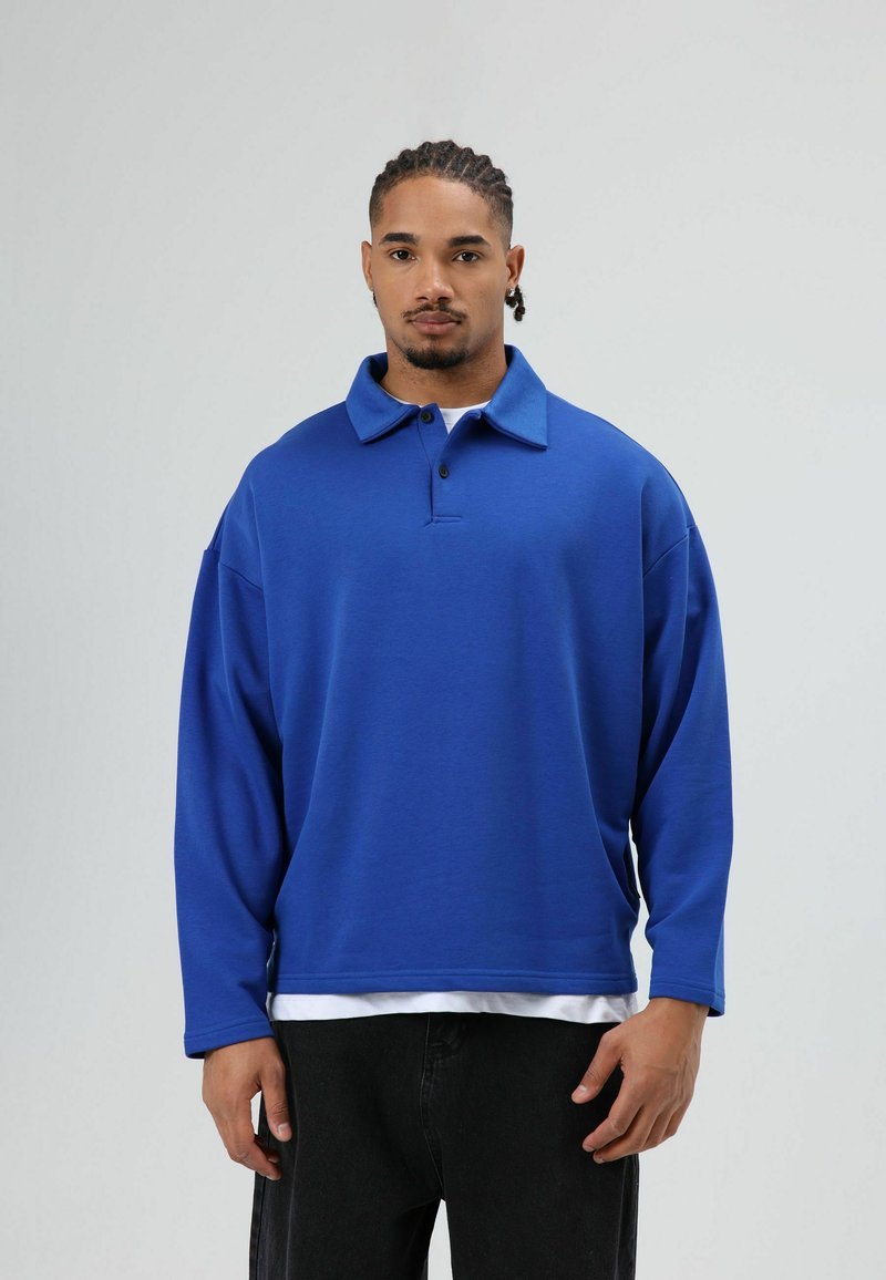 Blauw poloshirt met lange mouwen en een kraag, met een losse pasvorm en dikke stof, gedragen over een wit ondershirt en een zwarte spijkerbroek.