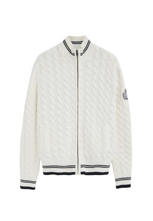 Jersey blanco de punto con cremallera y diseño de ochos, cuello, puños y bajo acanalados en rayas blancas y negras, y detalle de escudo en la manga izquierda.