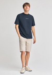 Mörkblå t-shirt med kort ärm med texten "POLO CLUB", tillsammans med beige shorts. Outfiten kompletteras av ljusa sneakers.