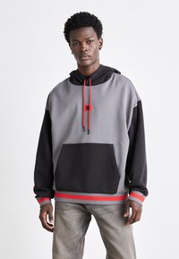 Haut de sweat à capuche en polaire gris et noir avec des accents rouges, capuche à cordon, poche avant et ourlet côtelé. Texture douce et coupe décontractée.