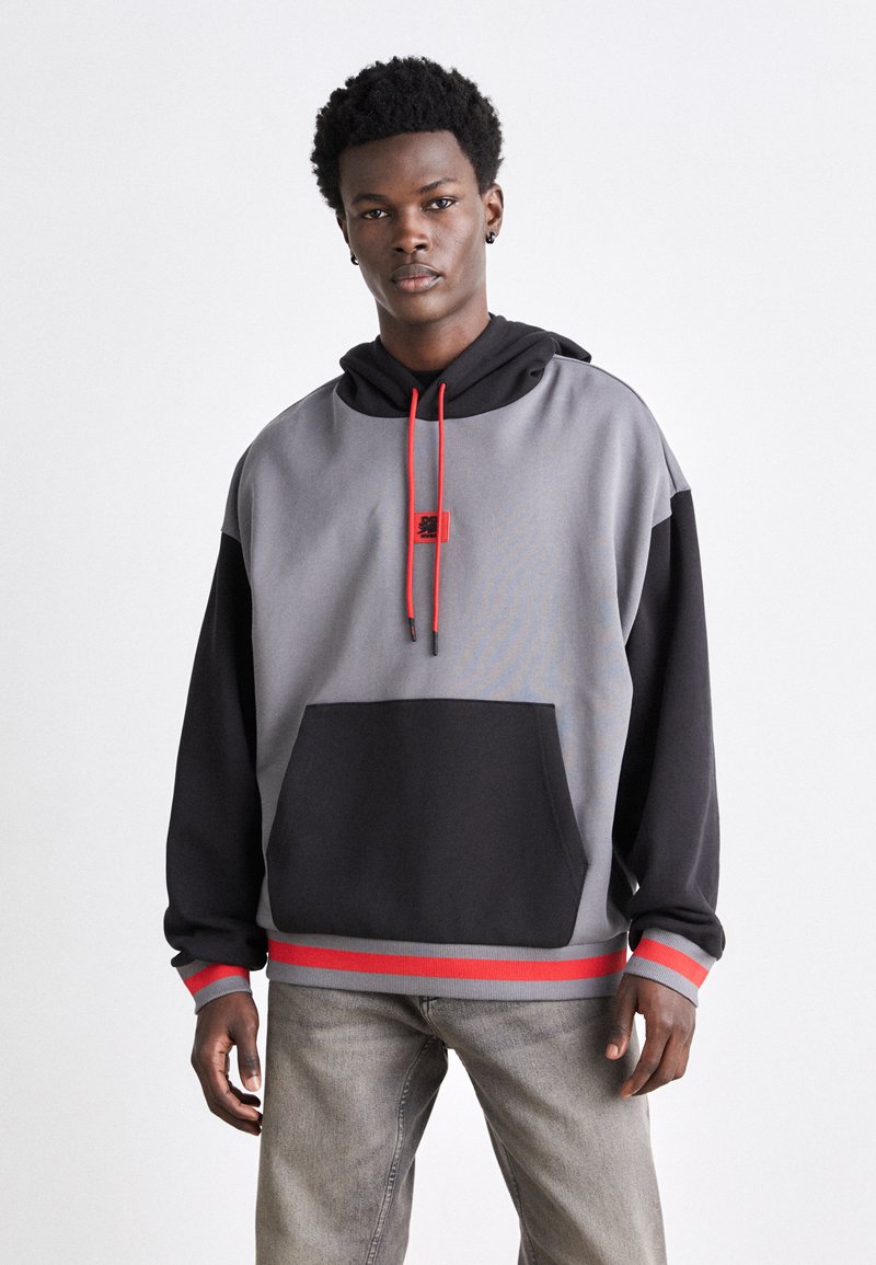 Haut de sweat à capuche en polaire gris et noir avec des accents rouges, capuche à cordon, poche avant et ourlet côtelé. Texture douce et coupe décontractée.