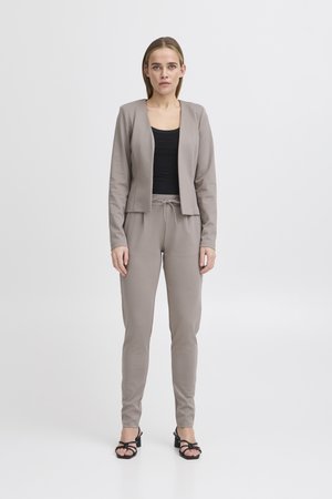 Femeie în picioare purtând un sacou taupe și pantaloni jogger asortați, cu un top negru și sandale cu toc negre, pe fundal alb.