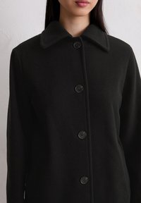 Cappotto in misto lana nera con ampio colletto, chiusura a bottoni e tessuto texturizzato. Presenta cinque bottoni neri rotondi lungo la parte anteriore.