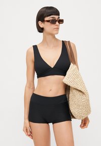 Ensemble de bikini noir côtelé comprenant un haut à col en V profond et un short taille haute. Le mannequin porte un sac en paille texturé et des lunettes de soleil en écaille.