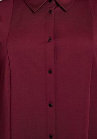 Close-up van een kastanjebruine shirt met zwarte drukknoopjes en een puntige kraag op een gestructureerde stofoppervlak.