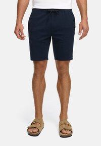 Homme portant un short bleu marine à cordon, une chemise blanche et des sandales beiges à deux lanières, se tenant sur un fond blanc.