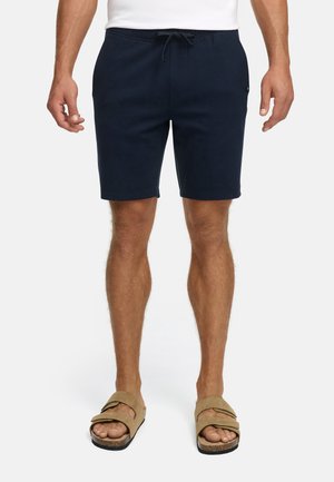 Mand iført navy blå snoretræk shorts, hvid skjorte og beige sandaler med dobbelte stropper, stående på en hvid baggrund.
