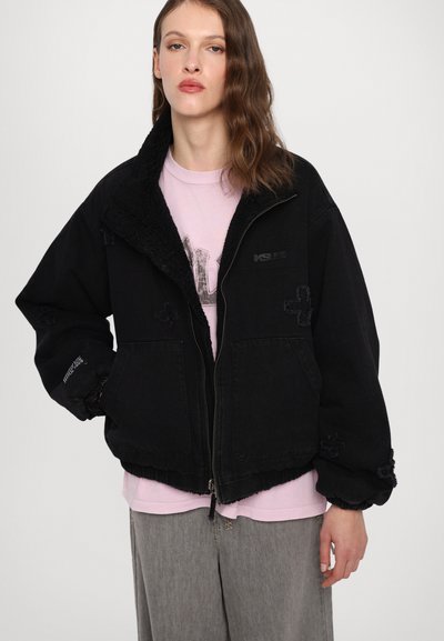 Black denim jacket with a textured, black fleece collar. Features front pockets, elastic cuffs, and subtle embroidered accents. Worn over a pink shirt.
