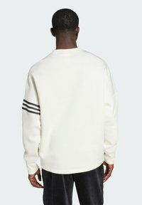 Sweatshirt blanc avec un col rond, des épaules tombantes et trois bandes noires sur la manche gauche. Texture lisse, coupe décontractée.