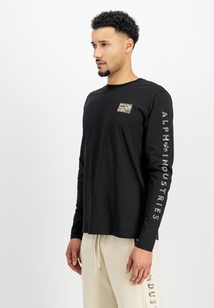 Crna majica dugih rukava s malim kamufliranim grbom na prsima i bijelim tekstom "ALPHA INDUSTRIES" duž desne rukave.