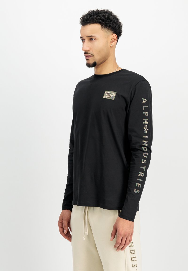 Camiseta negra de manga larga con un pequeño parche camuflado en el pecho y texto blanco "ALPHA INDUSTRIES" en la manga derecha.