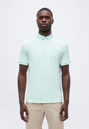 Joven hombre negro de pie, vistiendo una camiseta polo de manga corta color menta clara y pantalones beige, sobre un fondo blanco liso.