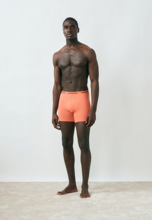 Muskelöser Mann steht barfuß vor einer schlichten Wand, trägt korallenfarbene Björn Borg Boxershorts und blickt entspannt nach vorne.