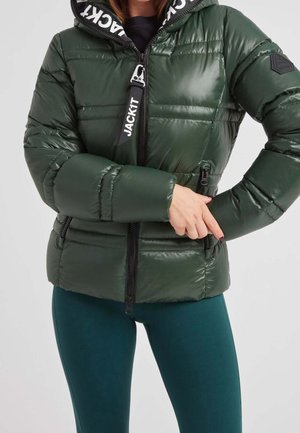 Femme portant une veste matelassée vert foncé avec une fermeture éclair visible et une étiquette avec logo, ajustant ou ouvrant la fermeture éclair de la poche latérale, montrée des épaules jusqu'aux hanches.