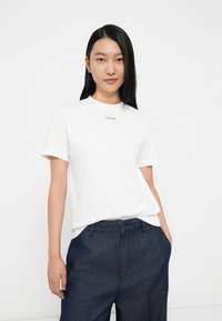 Valge puuvillane T-särk lühikeste varrukatega ja ümmarguse kaelusega, mille ees on väike must "Calvin Klein" logo. Kombineeritud tumedate teksapükstega.