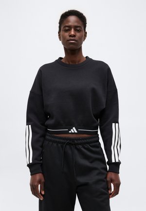 Persona vistiendo una sudadera negra recortada con rayas blancas en las mangas y pantalones negros con cordón, de pie frente a un fondo claro y liso.