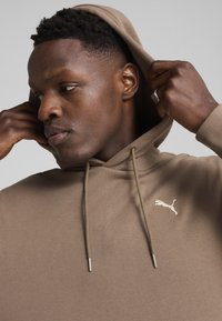 Sudadera marrón con cordones y pequeño logo blanco de Puma. Tejido suave y corte relajado. La capucha está retirada, mostrando las características faciales.