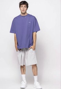 Lila, túméretes póló logóval a mellkason, világosszürke térdnadrággal párosítva. A megjelenést fehér tornacipő és bokazokni teszi teljessé.