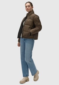 Bruin en zwart geblokt pufferjack met een hoge kraag, ritssluiting en zijzakken, gecombineerd met lichtblauwe jeans en beige platformlaarzen.