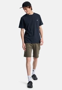 Camiseta de algodón negra con un pequeño logo, combinada con pantalones cortos de cargo verdes oliva, calcetines blancos y zuecos negros. Atuendo casual para el aire libre.