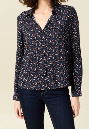 Vrouw die een marineblauw blouse met lange mouwen draagt, bedrukt met kleine roze en witte bloemenpatronen, gecombineerd met donkerblauwe spijkerbroek.