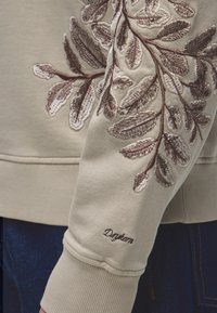 Manche de sweat beige avec des branches feuillues brodées en détail en marron et crème et un petit texte brodé en marron foncé près du poignet.