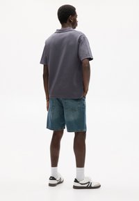 Gray polo shirt, denim shorts, white sneakers with black accents. Model stands with back turned, showcasing relaxed fit and casual style.