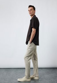 Mann steht mit den Händen in den Taschen, trägt ein schwarzes Kurzarmhemd, beige Hosen und hellbeige Sneakers auf grauem Boden.