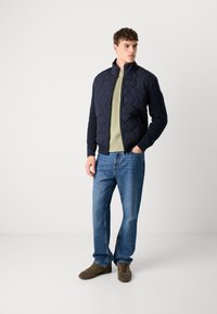 Marineblå quiltet jakke med rillede ærmer, parret med en lys grøn sweater og blå jeans. Model iført brune sko, står på en neutral baggrund.