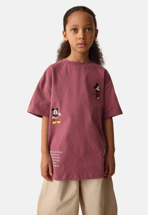 Barn med krøllet hår iført oversized vinrød T-shirt med Mickey Mouse-print og beige bukser med vide ben på ensfarvet baggrund.