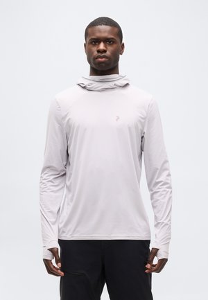 Homme portant un sweat à capuche gris clair à manches longues avec des trous pour les pouces et un pantalon noir, debout devant un fond blanc uni.