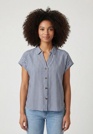 Donna con capelli ricci indossa una camicia a righe verticali blu e bianche a manica corta con bottoni e jeans azzurri, in piedi di fronte.