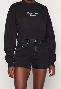 Svart kort sweatshirt med vit "Calvin Klein Jeans" logotyp, tillsammans med högmidjade svarta denimshorts med fransad nederkant.