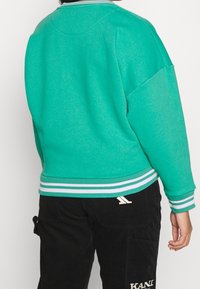 Persona che indossa un felpa color turchese con strisce bianche sui polsini e l'orlo, abbinata a pantaloni in corduroy neri con testo ricamato bianco.