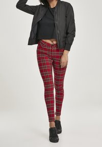 Veste bomber noire, design court ; legging à carreaux rouges avec accents noirs et verts ; modèle portant des baskets noires.