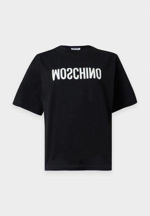 Kobieta w czarnym t-shircie Moschino, białych dżinsach z naszywkami w kształcie serca na kolanach i czarnych sandałach na obcasie, stoi z jedną ręką w kieszeni.