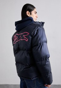 Veste puffer marine avec rembourrage épais, présentant un design graphique rose au dos. Superposée sur un hoodie bleu foncé, fabriqué dans un matériau brillant.