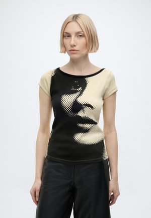 Femme aux cheveux courts et blonds portant un haut noir et beige à manches courtes avec un grand motif abstrait représentant un visage, associé à un pantalon noir.