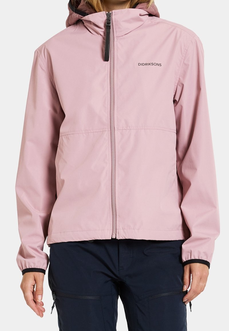 Femme portant une veste à capuche rose clair avec fermeture éclair à l'avant et logo, associée à un pantalon bleu marine foncé avec poches zippées.